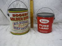 Front view of two vintage metal tins with metal handles and lids, one large cream colored Rogers Golden Syrup tin, one smaller red Burns Pure Lard tin with branding and some wear.