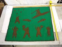 Full view of the green felt art panel with brown felt figures depicting traditional indigenous scenes including a paddler in a canoe, animals, and people.