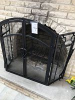 Front view of the black metal three-panel fireplace screen installed in a stone fireplace with fireplace tools visible on the side.