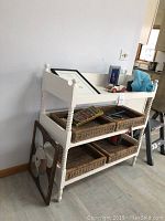 White painted wooden shelving unit with turned legs and tray top shelf, holding four wicker baskets on two lower shelves. Various small decorative items placed on top shelf.