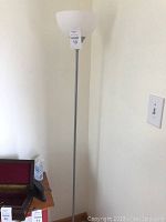 Full view of a single white glass shade floor lamp with silver metal pole and round base, positioned by a wall next to a side table with miscellaneous items.