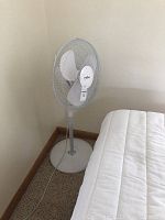 Front-left angle view of the white stand-up oscillating floor fan near a bed and wall.