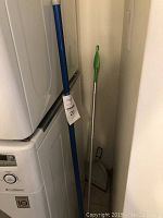 Photo of broom with blue handle, mop with green handle, and dustpan next to stacked washer/dryer units.