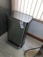 Closed stainless steel rectangular trash can with foot pedal and brushed finish, against a wall and beside a window with vertical blinds.
