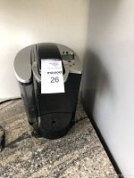 Front-angle photo of the black Keurig single-serve coffee maker on a countertop with seller tag 26 attached.