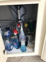 Items stored under kitchen sink including spray bottles, cleaning liquids, metal dish rack, and cleaning tools.