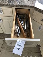 Photo of wooden drawer in kitchen containing stainless steel grilling tools with wooden handles, showing tongs, spatula, fork and other tools.