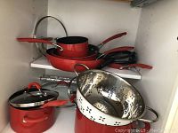 Cabinet interior with assorted red pots and pans, glass lids, and a stainless steel colander.