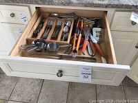 Top view of an open kitchen drawer displaying a wooden organizer tray containing a flatware set of forks, knives, and spoons; stainless steel measuring cups; various kitchen knives, tongs, and scissors with orange and black handles; a slotted spatula, and a wooden pestle.