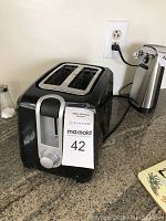 Photo of a black two-slice toaster on a kitchen counter, plugged into an outlet, with visible dial control and slots for bread.