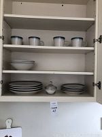 Open cabinet showing four cups on the top shelf, stack of bowls and different sized plates on lower shelves, and a glass sugar bowl at center bottom shelf.