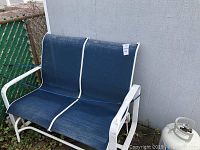 Two person lounge bench with white metal frame and blue fabric seating positioned outdoors next to a grey wall and a chain-link fence.