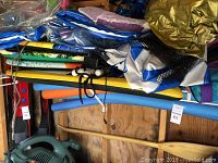 Photo showing assorted inflatable or fabric swim gear items, life jackets, foam kick boards, and pool noodles (not included). Items stacked on a wood shelf.