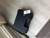 Black plastic Ryobi-branded hard carry case for drill set, closed and positioned on floor near doorway.