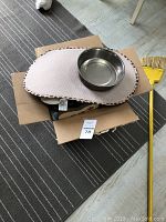 Photo showing a cardboard box containing a white oval pet feeding mat with brown speckled edges, and on top a round stainless steel pet bowl, placed on a striped rug floor.