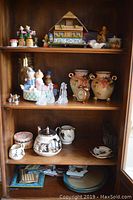 Wooden cabinet top three shelves with various porcelain, figurines, clock, glassware, and decorative items including Noritake urns, Telechron clock, and music box
