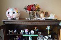 Shelf with pink piggy bank, colorful artificial flower arrangement in textured glass vase, assorted figurines including roosters, horses, and flower ceramics, and a teapot on a wooden shelf with decorative cloth.