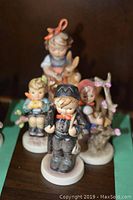 Four Goebel Hummel figurines arranged close, showing boy in black outfit at front and the other three figurines behind.