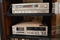 Shelf with two vintage audio units: top is a silver Technics FM/AM stereo receiver SA-222, bottom is a Technics stereo cassette deck M13 with cassette inserted, both show minor wear.