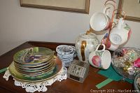Photo showing 4 porcelain fruit design mugs hanging on copper stand, piggy bank with gold handle and butterfly design, and stack of coasters on wooden surface.