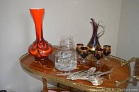 Photo of a tall orange-red crystal vase, crystal glasses, and a purple decanter set with gold overlay rims on a wooden bar cart.