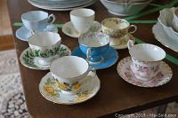 Photo showing seven assorted vintage tea cups and saucers of various floral patterns and colors arranged on a wooden surface, including yellow and pink roses, green ivy, and blue interiors.