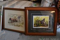 Two framed prints resting on a patterned surface: on left is a barn and silo scene, on right is a signed and numbered print of rabbit with flowers.