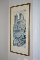 Framed print showing Notre Dame Cathedral along a riverside scene with artwork displays and boats, hung on wall