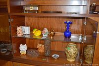 Display cabinet shelf with various ceramic animal piggy banks, animal figurines, small vases and plates.