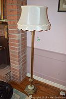 Full view of the floor lamp placed next to a brick fireplace and wall, showing overall height, metal base, column, and fabric lampshade.