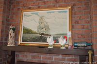 Mantel shelf with tall lighthouse ceramic, framed ship painting, two ceramic roosters, large German stein, and ship in bottle displayed on blue-green bottle with wooden stand.