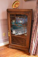 Full view of vintage wooden display cabinet standing against wall with glass door and decorative windowpane design. Scratches and minor wear visible.