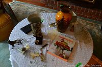 Photo showing various items on lace-covered table including SMF hand painted vase with tree scene, German Majolica vase, brandy glass with warmer, glass and metal candleholder, miniature glass collection, and a book titled 'Room Recipes'.
