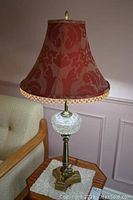 Full side view of the table lamp on a side table next to a couch, showing overall size, shape, and design including the red fabric lampshade with beaded fringe.
