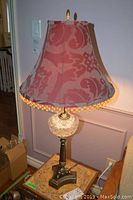 Full view of the table lamp showing metal base, glass decorative element, fabric shade, and finial on top.