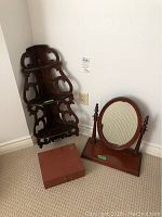 Photo showing wooden corner wall shelf, wooden box, and stand mirror placed near a white wall and electrical outlet.