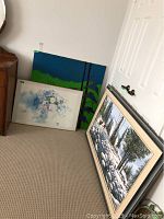 Three paintings leaning against a wall: a large winter forest oil painting with moose in frame, a floral image in a metal frame, and a colorful abstract painting.