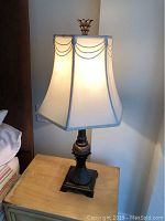 Full view of a lit table lamp showing ornate dark base with detailed pattern and hexagonal cream fabric shade with swag design, approximately 30 inches tall.