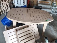 Photo showing beige resin rectangular outdoor table with slatted top, surrounded by several beige resin chairs, exhibiting the setting and style of the lot.