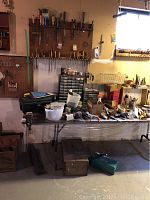 Wide view of folding table covered with hand tools on top and tools stored below. Wooden racks on wall hold chisels and files.