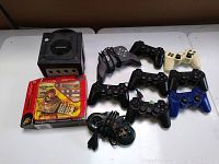 Image showing a black Nintendo GameCube console along with 9 PlayStation-style controllers of various colors and two arcade-style wired controllers, plus a boxed handheld LCD game.