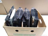 Photo showing four PlayStation 3 consoles inside a cardboard box viewed from top front angle, consoles standing vertically.