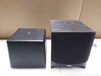 Two black powered subwoofers shown side by side, one with fabric front grille and wood grain finish, the other cube shaped with plain black finish