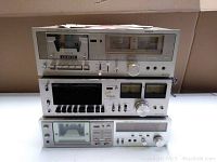 Lot of 3 vintage stereo cassette decks stacked, showing front panels including cassette compartments, analog VU meters, and control buttons.