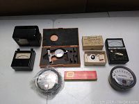 Overview of all items in the lot showing multiple vintage analog meters, dial indicator set in orange case, stylus pressure gauge in box, and round MAGNEHELIC gauge