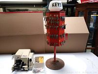 Key cutting machine positioned next to red three-sided rotary key blank carousel stand topped with a white illuminated 'KEYS' sign