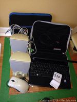 Photo showing MSI netbook, two beige speakers, white wired mouse, power cables, and two carrying cases on a floor with green tape borders.