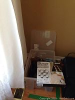 Plastic bin holding various outdoor solar lights and box of LED deck lights near curtain and vent on floor.