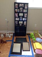 Photo showing the tall black wall-mounted curio display cabinet with various photos inside and several picture frames on the floor below it, next to plastic containers.