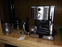 Wide shot of espresso machine, pewter mug, French press, and metal creamer pitcher on wooden shelf.